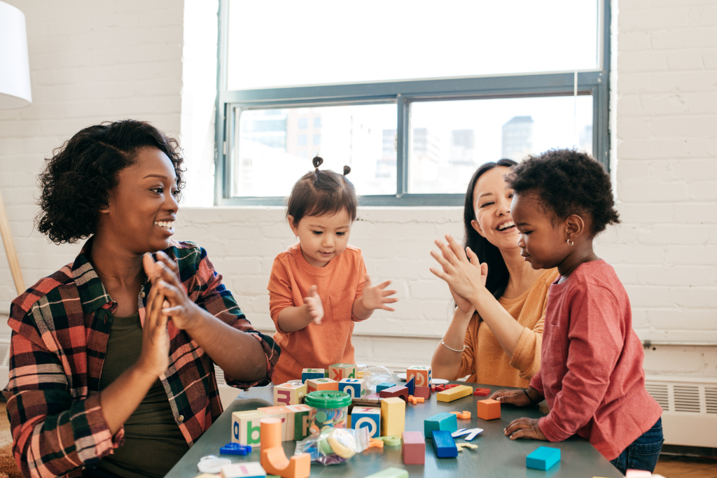Two women clapping hands with two toddlers at a table with building blocks.