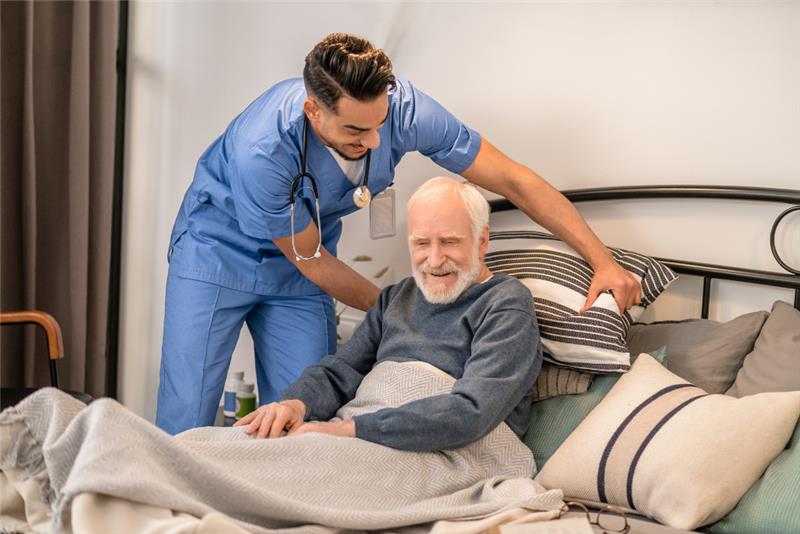 A young male caregiver in blue scrubs adjusts the pillow behind an elderly man sitting in a bed.
